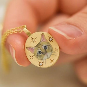 Könnte beinhalten: Goldfarbene Anhängerkette mit detaillierter Illustration eines Katzenkopfes. Der runde Anhänger ist mit Sternen und Diamanten verziert. Die Kette wird von einer Person gehalten.