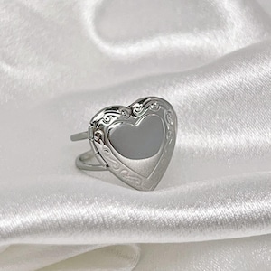 Könnte beinhalten: Silberfarbener herzförmiger Locket-Ring. Das Medaillon hat ein eingraviertes Design an den Rändern und eine glatte, reflektierende Oberfläche in der Mitte. Der Ring hat ein einfaches Doppelschlaufendesign.