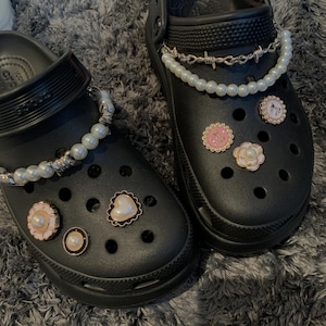 May include: A pair of black Crocs shoes decorated with pearl and silver-tone charms. The charms include a pearl chain, a heart-shaped charm, and flower-shaped charms with pink and white details. The shoes are shown on a grey, textured surface.