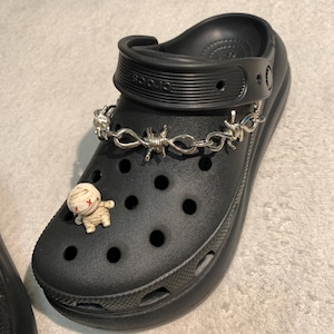 May include: Black clog shoe featuring a silver barbed wire chain and a small mummy charm. The shoe has multiple holes and a pivoting heel strap. The mummy charm is beige with red cross markings.