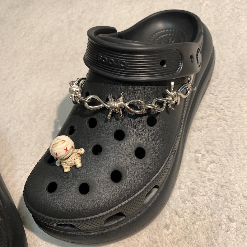 May include: Black clog shoe featuring a silver barbed wire chain and a small mummy charm. The shoe has multiple holes and a pivoting heel strap. The mummy charm is beige with red cross markings.