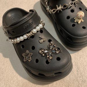 May include: Black clogs decorated with charms. One clog has a pearl and silver bracelet, a star, a butterfly, and cherry charms. The other clog features a barbed wire chain and a mummy charm. The clogs are on a light-coloured surface.