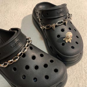 May include: Black platform clogs with silver chain and spike detailing. One clog has a small mummy charm. The clogs have multiple ventilation holes and a textured sole. The shoes are on a light grey surface.