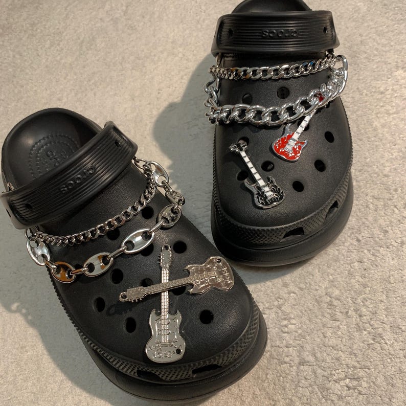 May include: Two black Crocs, each adorned with silver chains and guitar-themed charms. One shoe has a red and white guitar charm, the other has two silver guitar charms. The shoes are displayed on a light grey surface.