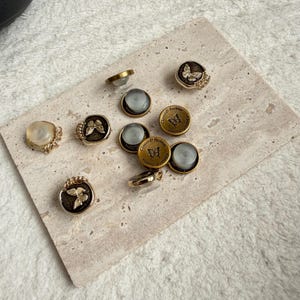 May include: A collection of decorative buttons with butterfly and floral designs. Some buttons are gold-toned with a butterfly motif, while others have a clear, iridescent center. Several buttons feature the text "Classics for Fashion".