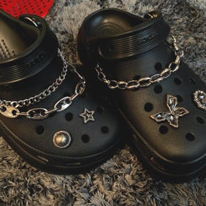 May include: Two black clogs embellished with silver chain details and decorative charms. One clog has a star-shaped charm and a round stud, the other a butterfly charm and a heart-shaped gem. The clogs are displayed on a grey surface.