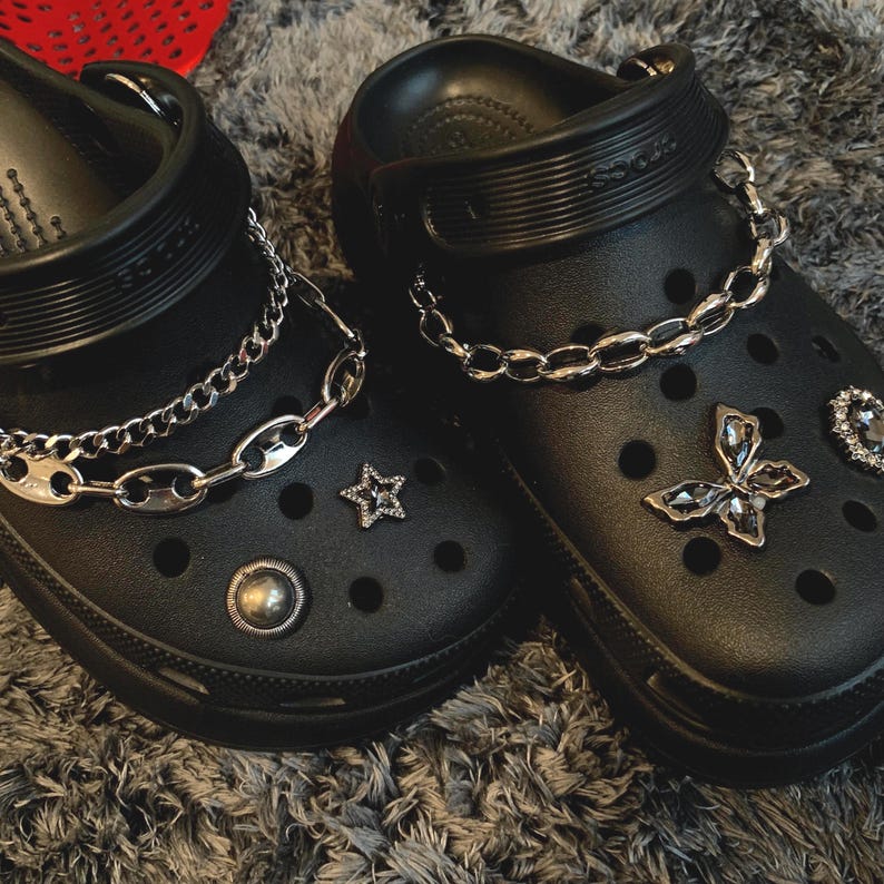 May include: Two black clogs embellished with silver chain details and decorative charms. One clog has a star-shaped charm and a round stud, the other a butterfly charm and a heart-shaped gem. The clogs are displayed on a grey surface.