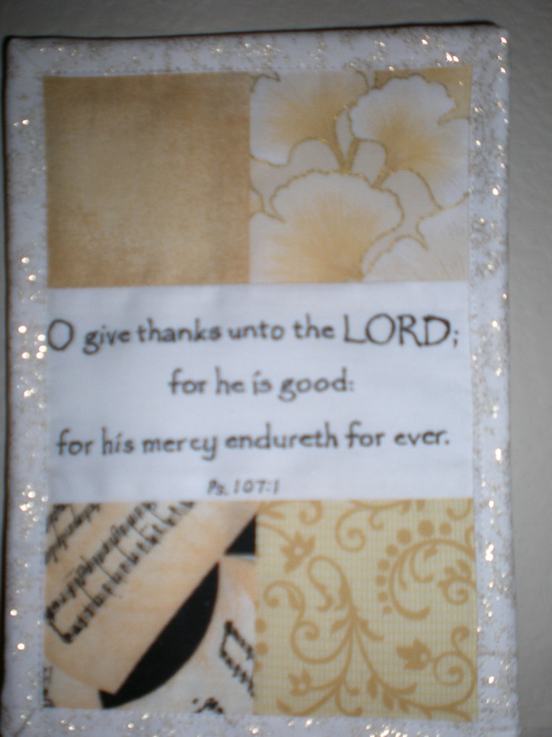 Puede incluir: Un cuadrado de tela blanca con un borde dorado y un patr&oacute;n floral dorado y blanco. El cuadrado de tela tiene un cuadrado de papel blanco con un vers&iacute;culo b&iacute;blico impreso en &eacute;l. El vers&iacute;culo dice "O give thanks unto the LORD; for he is good: for his mercy endureth for ever. Ps. 107:1".