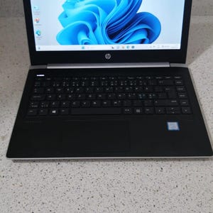以下が含まれることがあります： 画面に青い抽象的なデザインが施された、銀と黒のHP製ノートパソコン。ノートパソコンが開いており、キーボードが見えています。