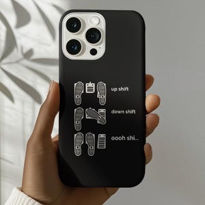 Puede incluir: Funda de teléfono negra con texto y gráficos blancos. El texto dice "up shift", "down shift" y "oooh shi...". Los gráficos son huellas de zapatos estilizadas y pedales de coche. La funda del teléfono se sostiene en una mano.