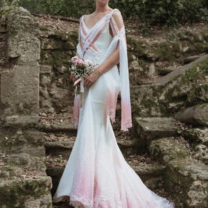Puede incluir: Un vestido de novia blanco con un efecto degradado rosa y detalles de encaje. El vestido tiene un corpiño ajustado y una falda fluida. La novia lleva un ramo de flores blancas y rosas.