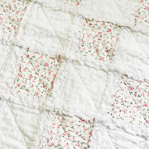 Könnte beinhalten: Eine weiße Steppdecke mit Patchwork-Design. Die Steppdecke hat Quadrate aus weißem Stoff und Quadrate aus Stoff mit einem rosa Blumenmuster. Die Ränder der floralen Quadrate sind mit gerüschtem weißem Stoff besetzt.