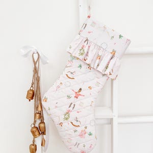 Peut inclure: Une chaussette de Noël blanche matelassée ornée d'un motif répétitif de casse-noisettes, de chevaux à bascule et de gui. Elle présente un revers à volants et une boucle de suspension. Une cordelette en jute avec des clochettes dorées.