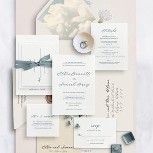 Puede incluir: Elegante juego de invitaciones de boda con un esquema de color crema y blanco. Incluye tarjetas de invitación, tarjetas RSVP, tarjetas de detalles y sobres. Una cinta azul y un pequeño plato con anillos añaden elementos decorativos. El sobre tiene un diseño de nubes.