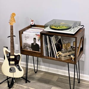 May include: A dark wood record player stand with black hairpin legs. The stand holds vinyl records, a turntable, and decorative items. A white electric guitar stands to the left.