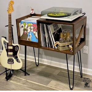 May include: A dark wood record player stand with black hairpin legs. The stand holds vinyl records and a turntable. A cream-colored electric guitar is on a stand to the left.