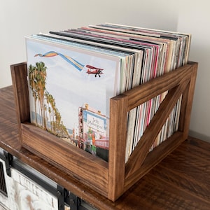 May include: A wooden record holder with a dark stain finish, holding a collection of vinyl records. The front record cover features a colorful image of palm trees, a biplane, and the text "Jacks Mannequin". The holder has a diagonal brace design.