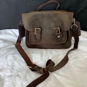 May include: A brown leather satchel bag with a long adjustable strap. The bag has two buckles on the front and a small pocket on the side.