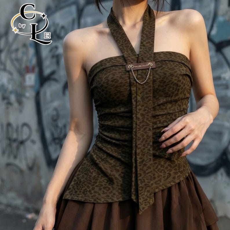 Puede incluir: Un top halter con estampado de leopardo en marr&oacute;n oscuro que presenta un cuerpo fruncido y una tira de tela vertical central adornada con un peque&ntilde;o parche rectangular marr&oacute;n y una fina cadena de metal plateado. Se combina con una falda marr&oacute;n.