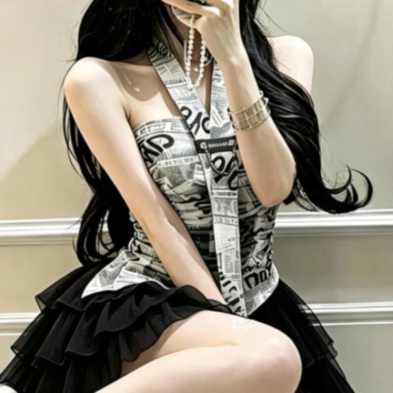 Puede incluir: Un top estilo halter con un patr&oacute;n de estampado de peri&oacute;dico en blanco y negro y una tira vertical central, combinado con una falda negra de volantes superpuestos. El top tiene un dise&ntilde;o ajustado y la falda consta de varios niveles de tela.