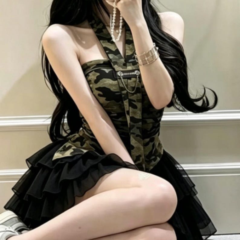 Puede incluir: Un top estilo halter con estampado de camuflaje en verde, marr&oacute;n y negro, con un detalle de cadena de metal plateado en el pecho, combinado con una falda de tul negro en capas con m&uacute;ltiples volantes que crean una silueta voluminosa.
