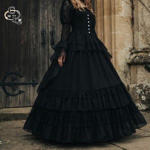 Puede incluir: Un vestido negro largo hasta el suelo con mangas abullonadas de encaje transparente, escote cuadrado y una fila vertical de botones blancos en el corpiño. La falda presenta múltiples capas escalonadas de tela y encaje para dar volumen.