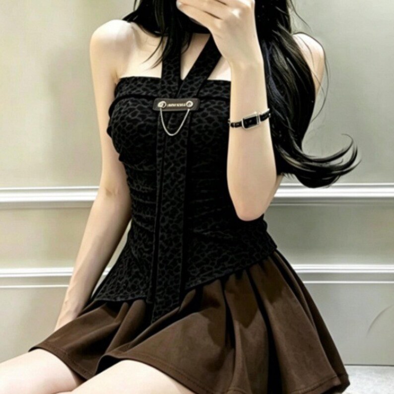 Puede incluir: Un top negro tipo halter con un sutil estampado animal, una tira central vertical de tela y un detalle de cadena de metal plateado sujeto a una leng&uuml;eta rectangular. El top se combina con una minifalda marr&oacute;n lisa, plisada y acampanada.
