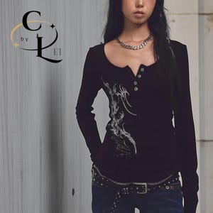 Pode incluir: Camisa preta de manga comprida com decote redondo e três botões. Apresenta um design tribal branco na frente. O logotipo "C by L E1" está no canto superior esquerdo.