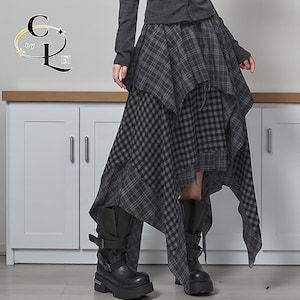 Puede incluir: Una falda asimétrica de varias capas en cuadros grises y negros. La falda tiene un diseño único y fluido con un dobladillo deshilachado. La modelo lleva botas de plataforma negras. El logotipo "C by L" es visible en la esquina superior izquierda.