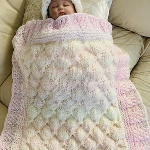 May include: A baby wrapped in a handmade, knitted blanket and hat. The blanket is white and pink, with a textured diamond pattern. The hat is white, and the blanket has a decorative edge. The baby is lying on a beige surface.