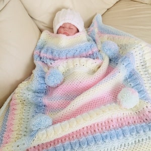May include: A baby swaddled in a pastel rainbow striped crocheted blanket with white pom-poms. The blanket has a crocheted border with a textured pattern.