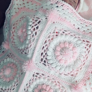 May include: A white and pink crocheted baby blanket with a delicate floral pattern and pom-poms. The blanket is made with soft yarn and is perfect for a newborn.