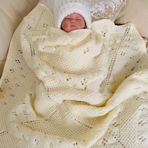 May include: A white crocheted baby blanket with a delicate lace pattern. The blanket is folded around a sleeping baby wearing a white crocheted hat.