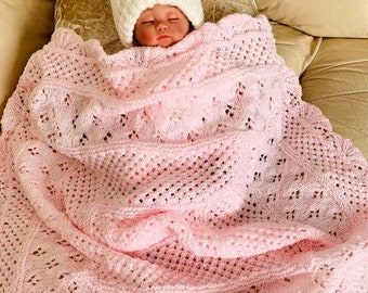 May include: A pink crocheted baby blanket with a delicate lace pattern. The blanket is folded and draped over a baby wearing a white knitted hat.