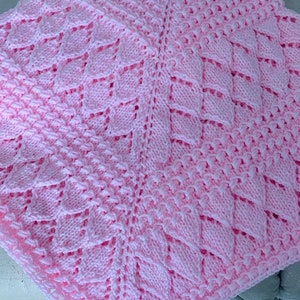 Op de afbeelding: Een roze gebreide babydeken met een delicaat, kantpatroon. De deken is gemaakt van zacht garen en heeft een delicate, vrouwelijke uitstraling.