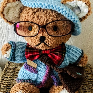 Peut inclure: Ours en peluche marron fait à la main, portant un béret bleu clair tricoté, des lunettes et un nœud papillon à carreaux. L'ours a une veste bleue assortie et une sacoche marron. L'ours est assis sur un panier tressé.