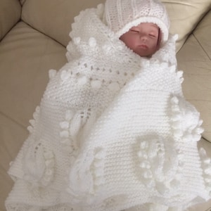 Puede incluir: Una manta de bebé de ganchillo blanco con un gorro de ganchillo blanco. La manta tiene un diseño texturizado con pompones en relieve.