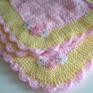 May include: A handmade, knitted baby blanket in soft pastel colors. The blanket features a pink and yellow design with a scalloped edge and decorative flower accents. The blanket is made of soft yarn, perfect for a baby.