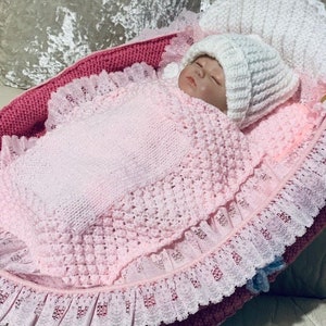 May include: A pink crocheted baby blanket with a white crocheted hat. The blanket has a lace trim and is in a pink and white basket.