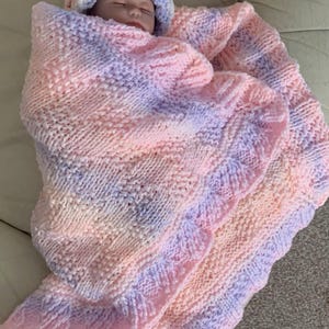 May include: A baby wrapped in a soft, hand-knitted blanket and matching hat. The blanket and hat are in shades of pink, lavender, and peach. The knitted blanket has a scalloped edge and a textured pattern. The hat has a pom-pom on top.