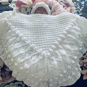 Può includere: Un neonato avvolto in una coperta bianca lavorata a maglia con un motivo a foglie e pompon strutturato. Il bambino indossa un cappello bianco lavorato a maglia. È visibile un giocattolo a forma di coniglio rosa e bianco lavorato a maglia.