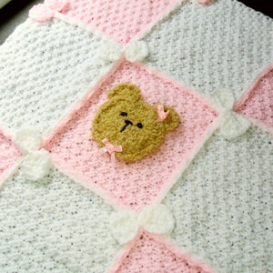 May include: A white and pink crocheted baby blanket with a brown teddy bear applique in the centre.