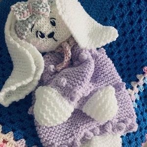 May include: A knitted lavender bunny with white accents and a pink bow. The bunny is lying on a blue and pink crocheted blanket.