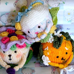May include: Three knitted stuffed toys in the shape of a flower, a pumpkin, and a person. The flower is purple and pink with yellow and purple flowers. The pumpkin is orange with green leaves. The person is white with pink and blue stripes.