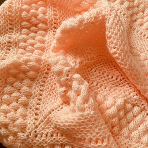 May include: A peach-coloured crocheted baby blanket with a textured pattern. The blanket is made with a soft yarn and has a delicate, feminine look.