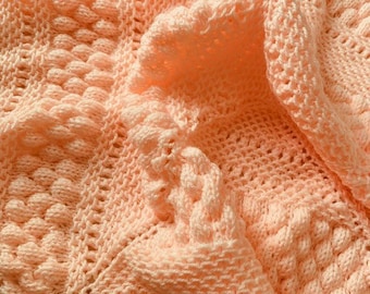 May include: A peach-colored crocheted baby blanket with a textured pattern. The blanket is made with a soft yarn and has a delicate, feminine look.