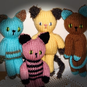 May include: Four knitted cat toys in different colours and patterns. The cats are blue, yellow, pink, and brown. They have black eyes and noses.