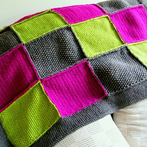 Può includere: Una coperta di maglia con un design a patchwork in tonalità di grigio, verde e rosa.