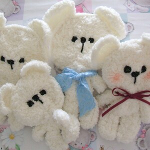 May include: Five fluffy white teddy bears with black embroidered facial features and small ears. One bear has a blue bow, and another has a burgundy bow. The bears are arranged on a patterned background.