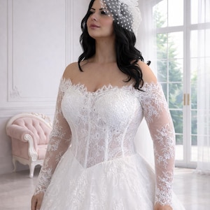 Peut inclure: Une robe de mari&eacute;e blanche &agrave; &eacute;paules d&eacute;nud&eacute;es avec des manches longues en dentelle, un corsage style corset aux d&eacute;tails floraux complexes et une jupe ample. Un bibi blanc assorti avec un voile &agrave; pois est port&eacute; sur la t&ecirc;te.